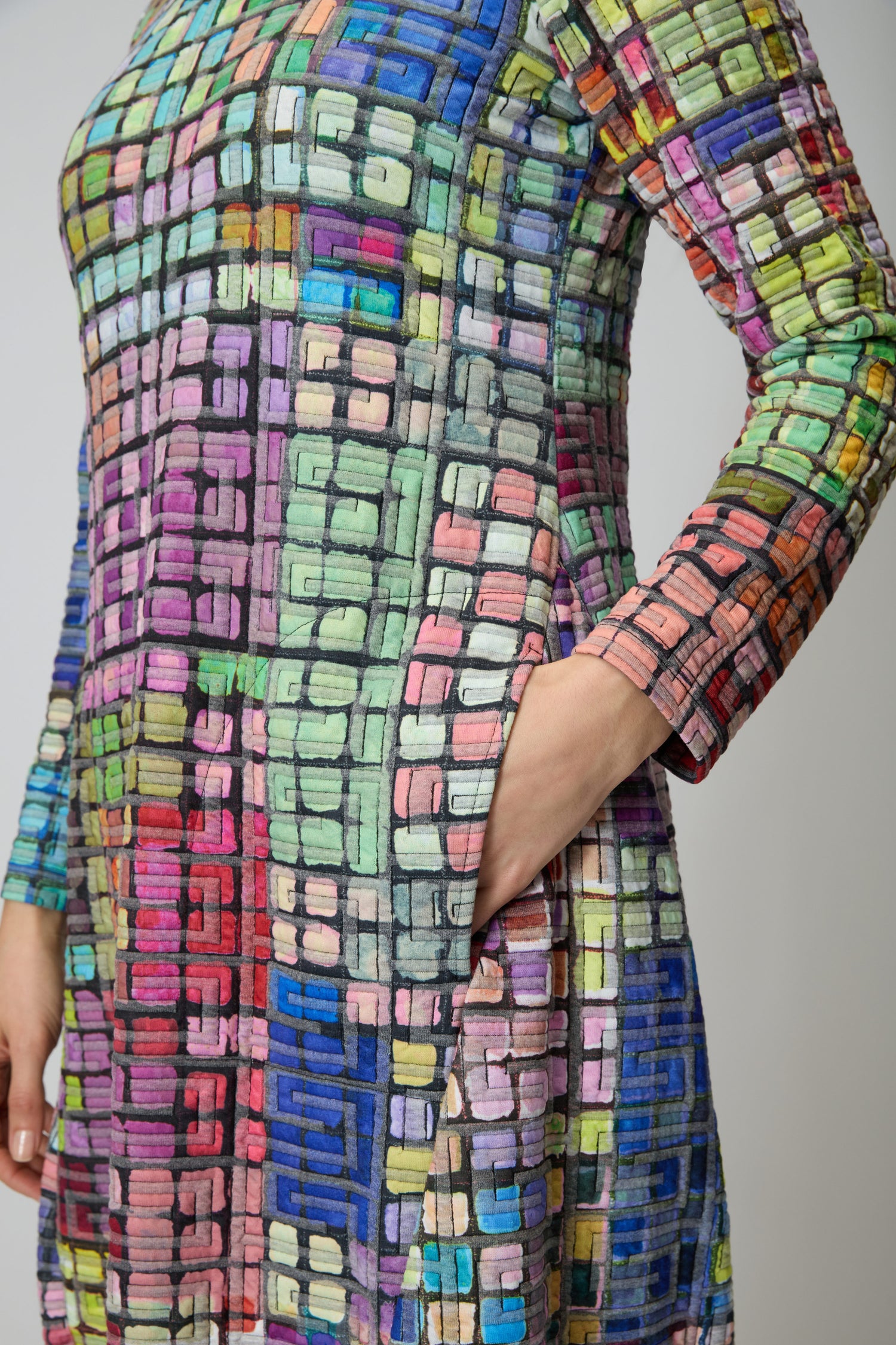 A person wears the Coloured Glass Tiles Jersey Bubble Dress, a long-sleeved bubble silhouette with a colorful geometric grid pattern, shown from shoulder to mid-thigh, one hand casually placed in a pocket.