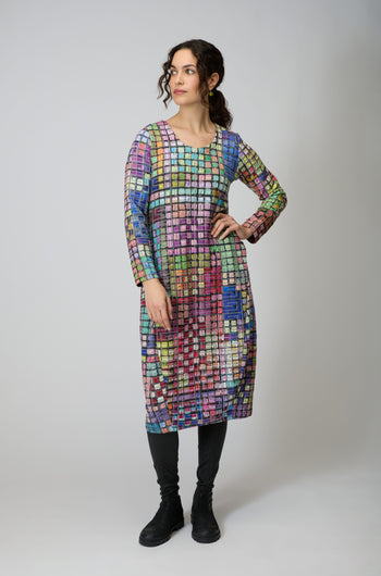 A woman faces slightly left, wearing the Coloured Glass Tiles Jersey Bubble Dress with a vibrant mosaic pattern, paired with black leggings and shoes, set against a neutral background.