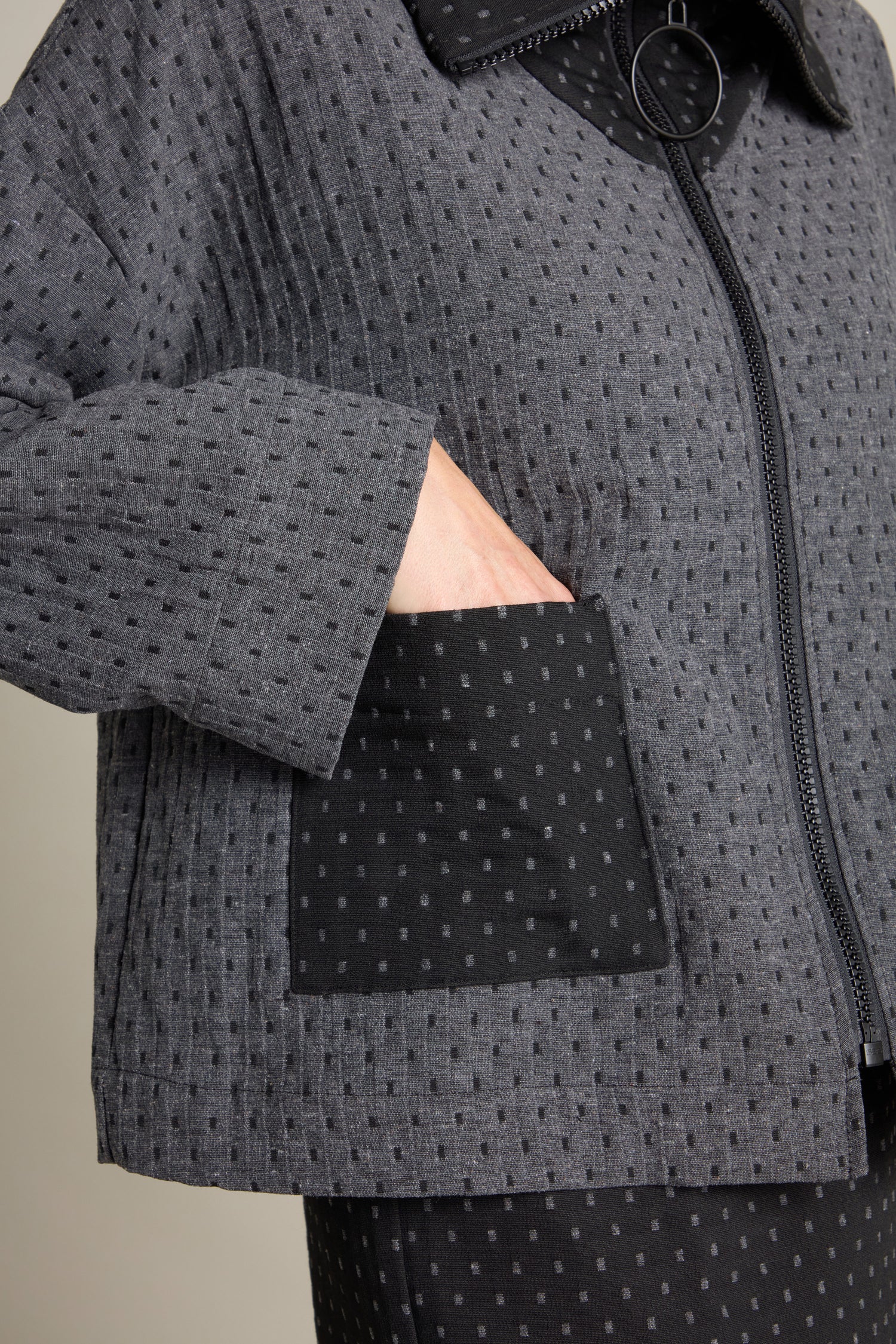 Close-up of a person wearing the Square Jacquard Boxy Jacket with a black polka-dot pattern, their left hand in a front pocket. The contemporary style jacket features a zipper and contrasting black pocket, showcasing its textured boxy fit.