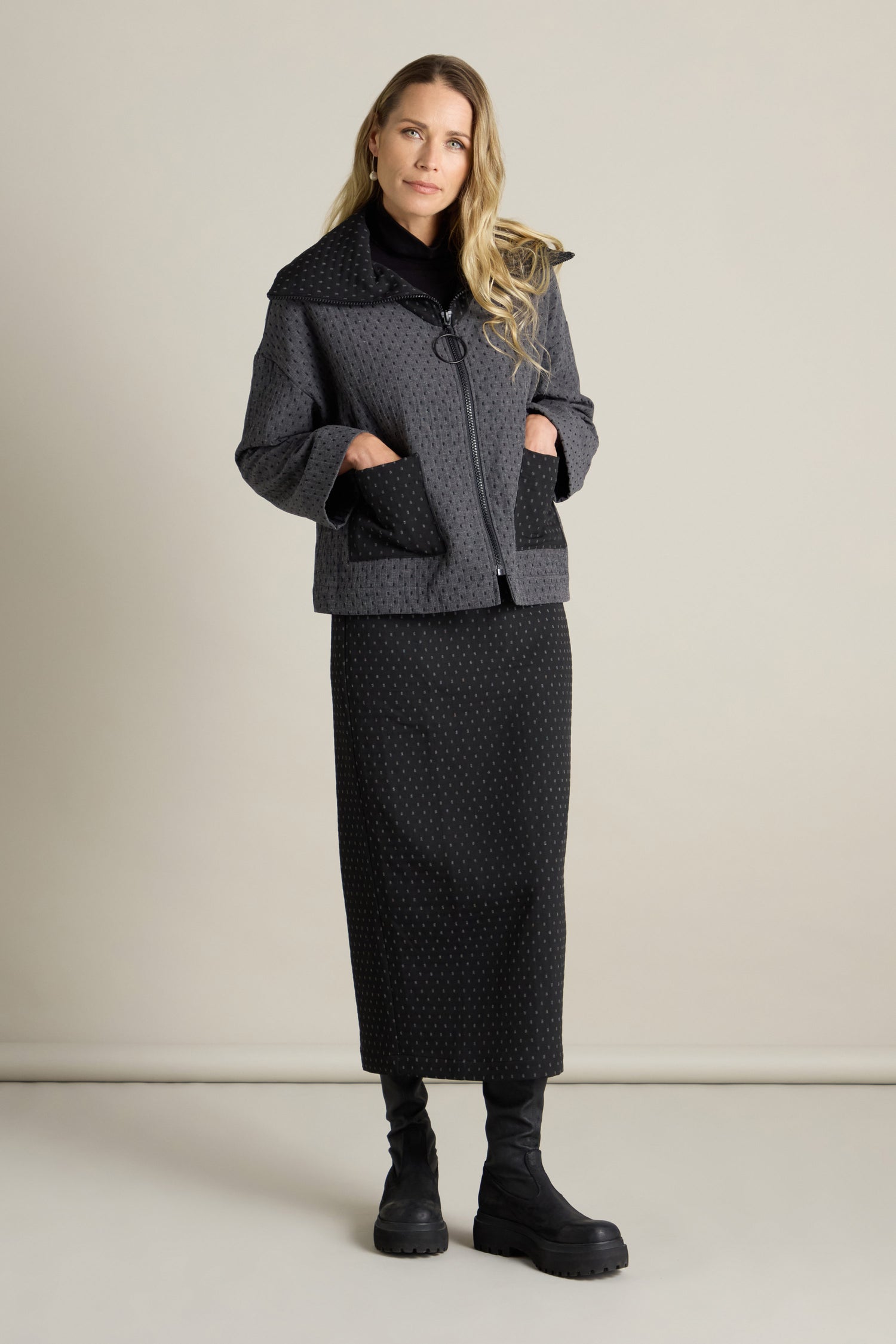 A person stands against a plain background, wearing a contemporary Square Jacquard Boxy Jacket in dark grey, featuring large pockets. They pair it with a matching long skirt and black boots to complete the outfit.