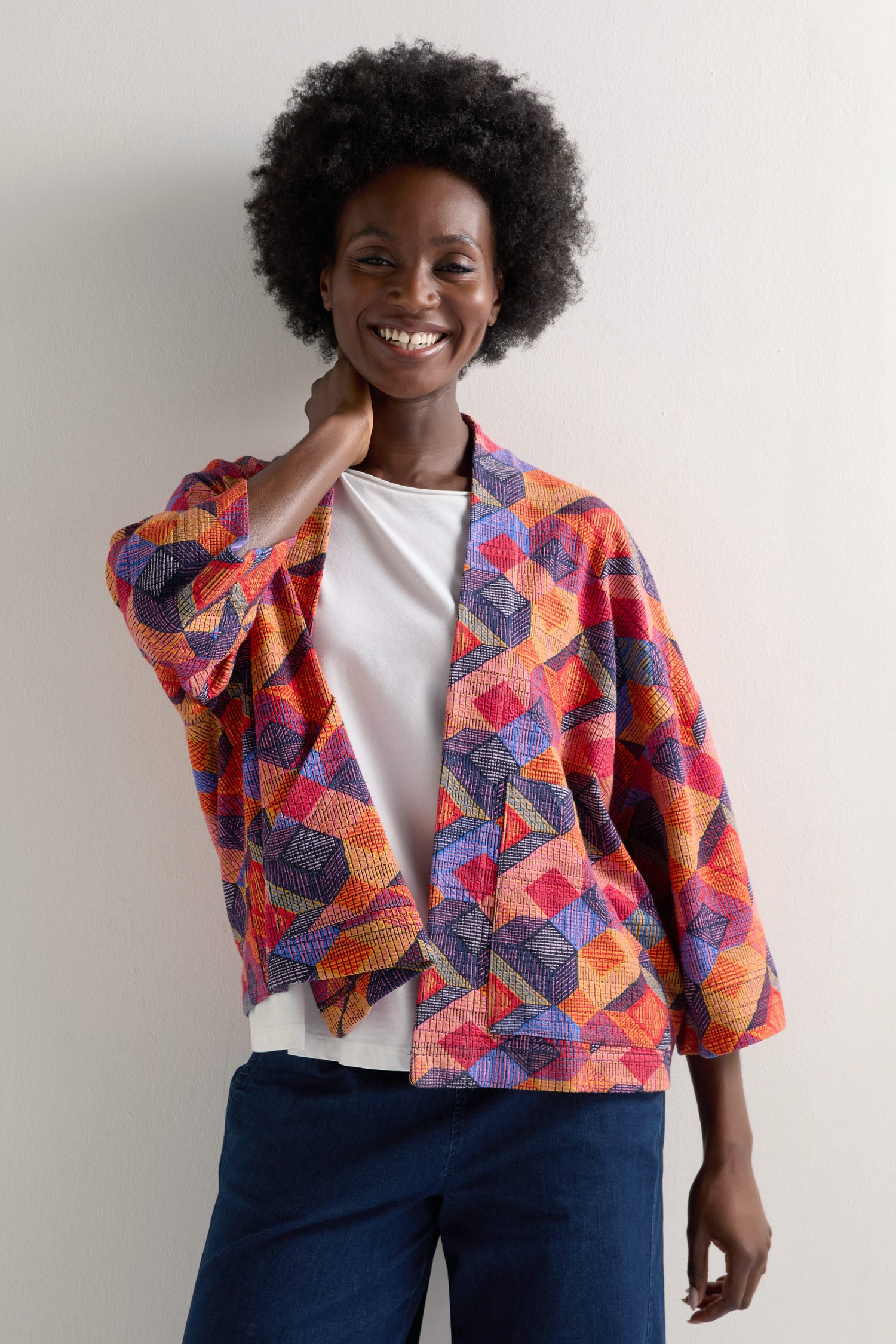 A woman smiles against a plain background, wearing the Bold Cubist Print Jacket in a contemporary style over a white top and blue pants.