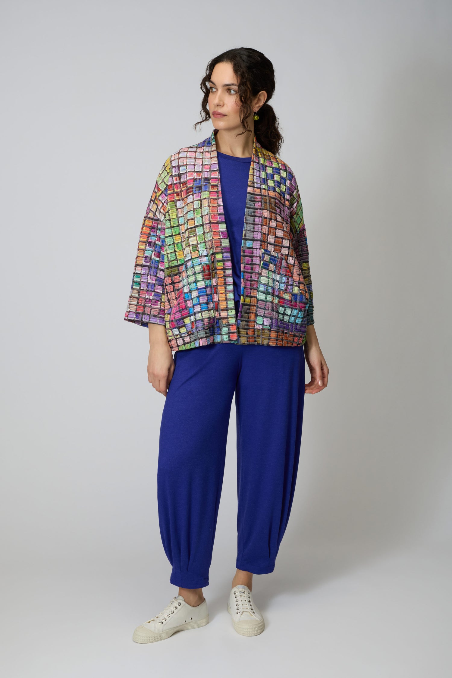 A woman wears the Coloured Glass Tiles Jersey Jacket—a lightweight blue top and pants set with vibrant glass tile patterns—paired with white sneakers against a plain light background.