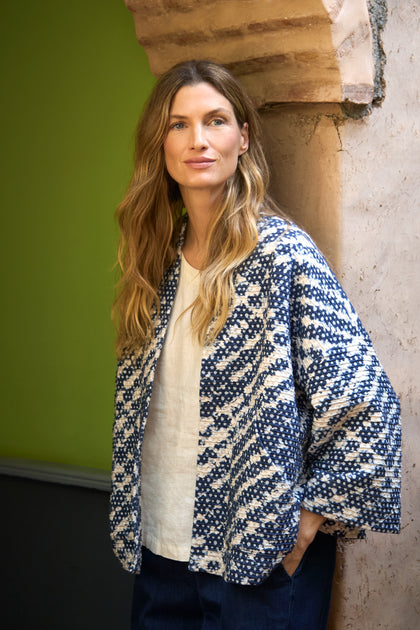 A woman with long blond hair stands by a stone arch, wearing a Diamond Tie Dye Jersey Jacket over a white shirt and looking at the camera.
