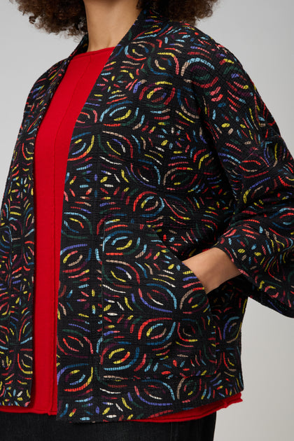 A person wearing a red top and the Mosaic Jacquard Jersey Jacket with colorful embroidered patterns stands with one hand in their pocket.