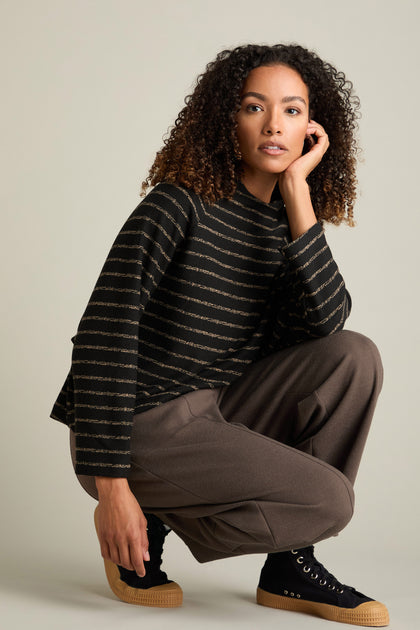 A person wearing a black and striped sweater paired with Marl Jersey Bubble Trousers in brown is squatting against a neutral background, embodying contemporary style.