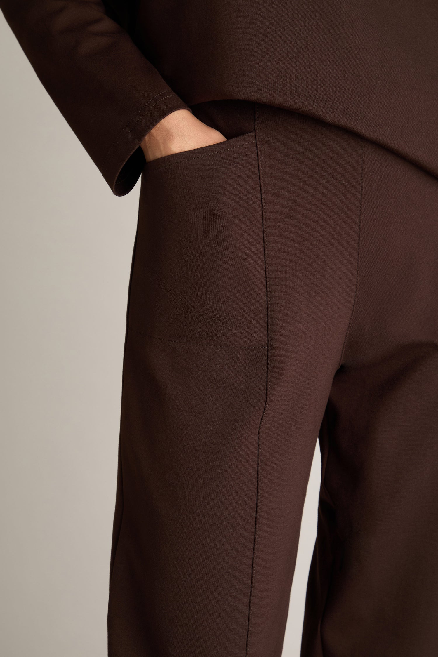 A close-up of a person’s waist showcases a hand in the side pocket of brown Ponte Bubble Trousers, emphasizing their flattering fit and elasticated back, paired with part of a coordinating brown top.