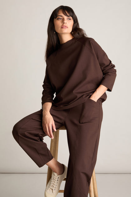 A woman wearing a flattering matching top and elasticated back Ponte Bubble Trouser in brown sits on a wooden stool, posing with one hand in her pocket against a plain background.