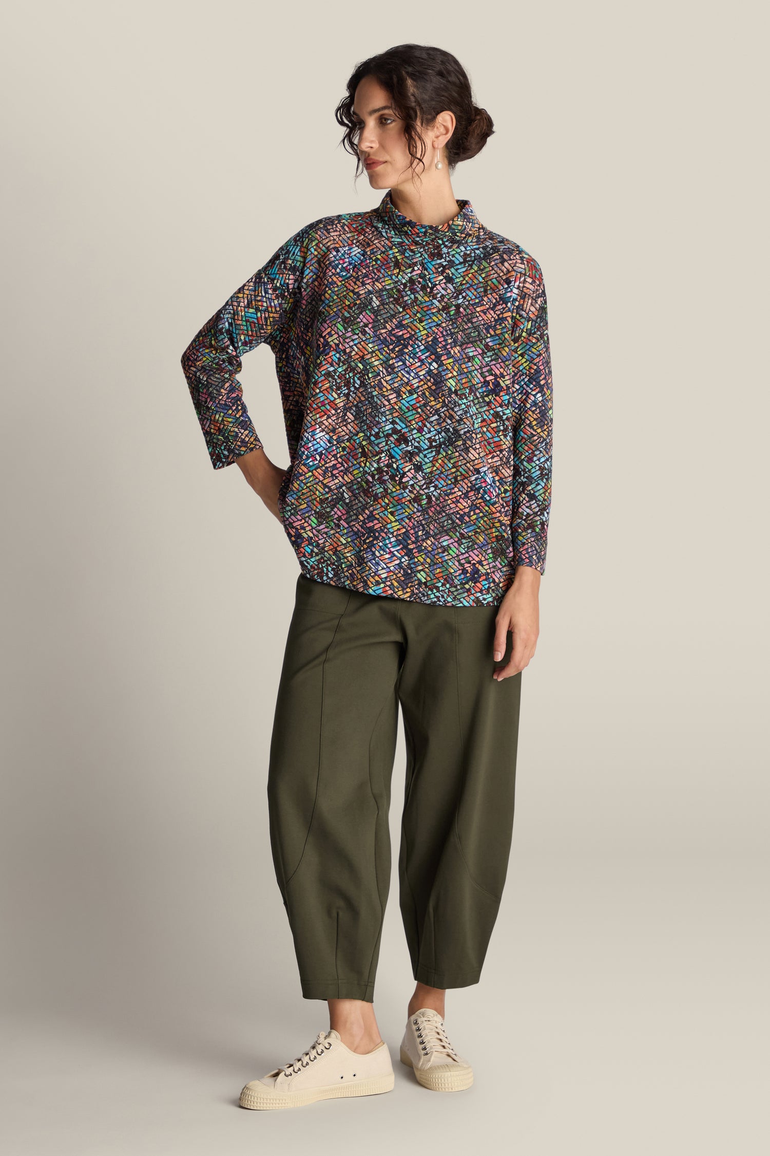 A woman stands against a neutral background wearing a multicolored patterned top, the Ponte Bubble Trouser in green made from mid-weight Ponte fabric, and white sneakers. She has her right hand resting on her hip, showcasing an outfit that combines comfort and fashion—a true year-round style staple.