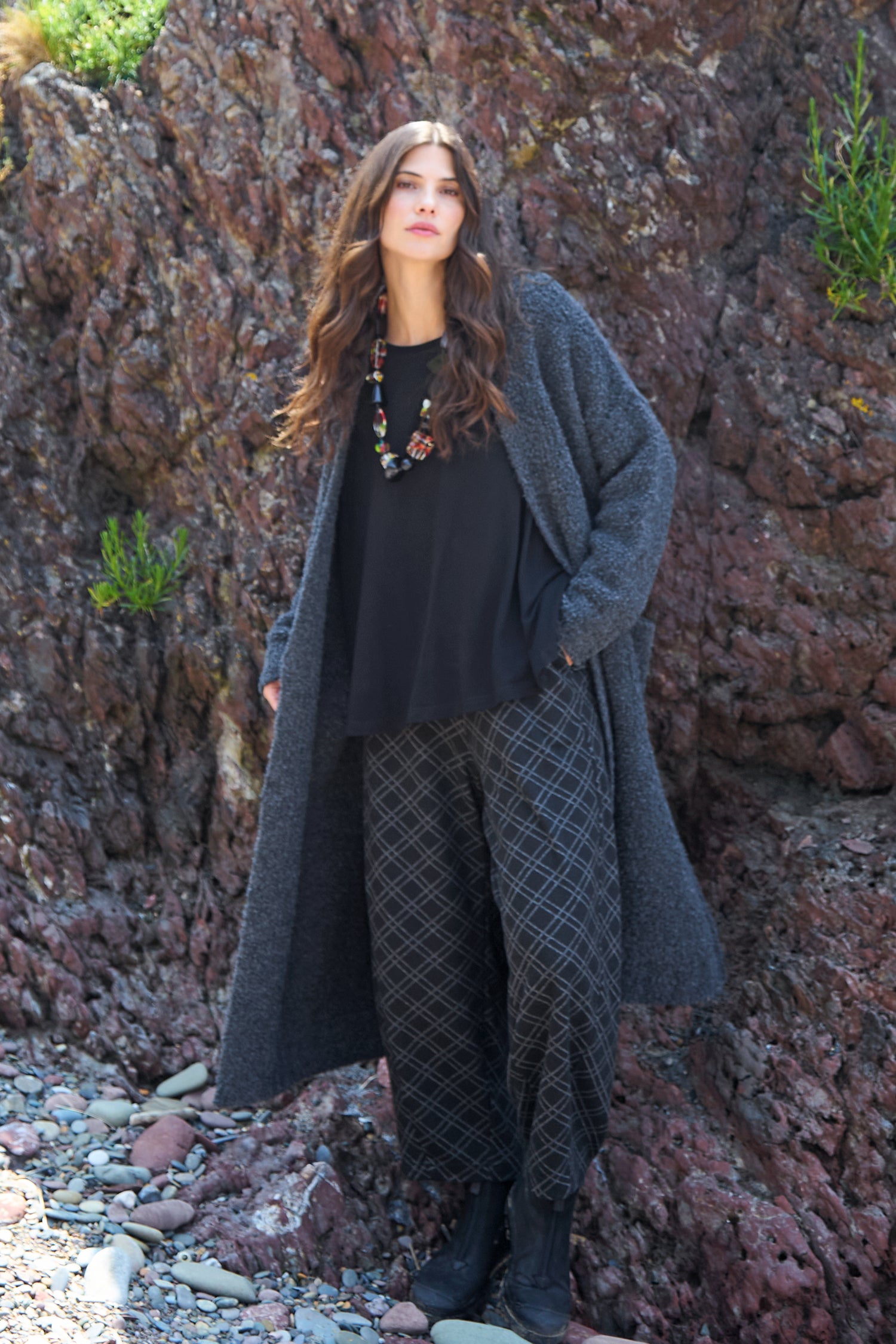 A woman stands on rocky ground in a long gray coat, black top, Square Jacquard Jersey Trousers with a grid pattern, black boots, and a colorful necklace, with a rocky cliff behind her.