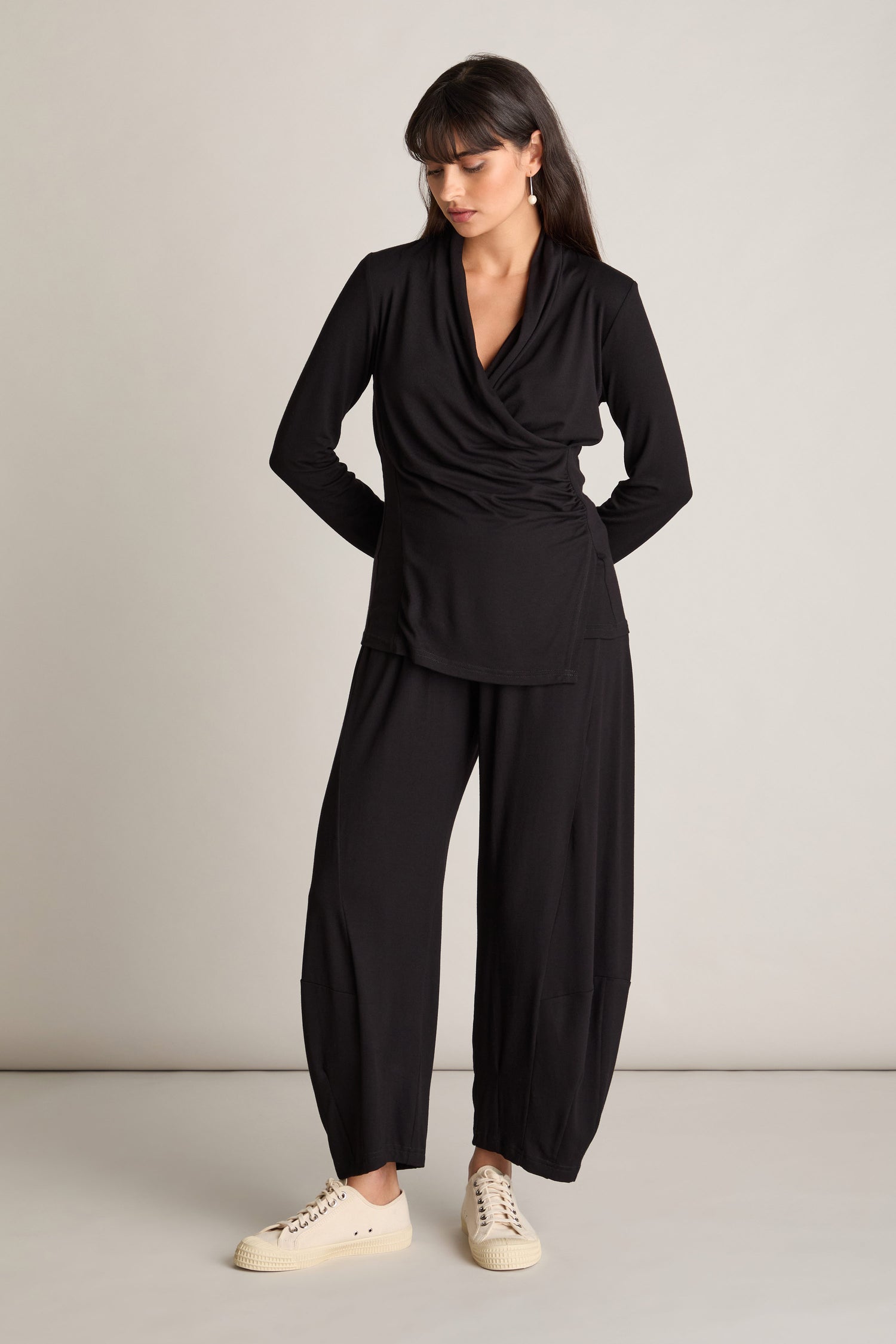 A woman stands against a plain background, wearing a black long-sleeve wrap top and matching black Fluid Crepe Bubble Trousers with white sneakers. Featuring an elasticated waist, she looks down to her right with her hands tucked in her pants pockets, exuding a chic co-ord look.