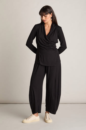 A woman stands against a plain background, wearing a black long-sleeve wrap top and matching black Fluid Crepe Bubble Trousers with white sneakers. Featuring an elasticated waist, she looks down to her right with her hands tucked in her pants pockets, exuding a chic co-ord look.