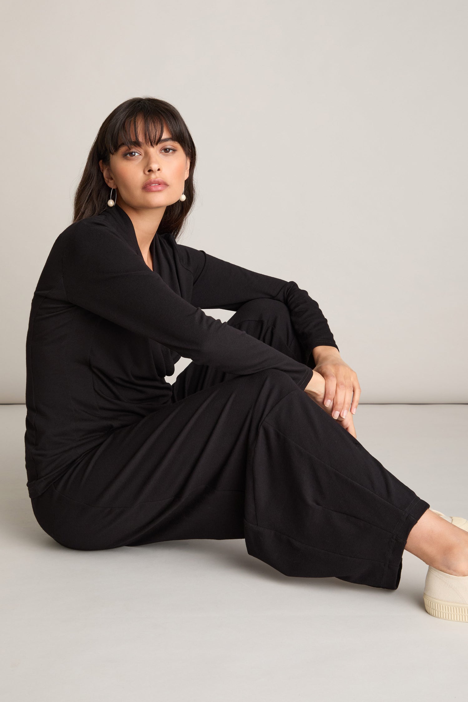 A person with long dark hair and bangs is seated on the floor against a plain background. They are donning a chic co-ord look featuring a black long-sleeved top and Fluid Crepe Bubble Trouser with an elasticated waist, paired with light-colored shoes.