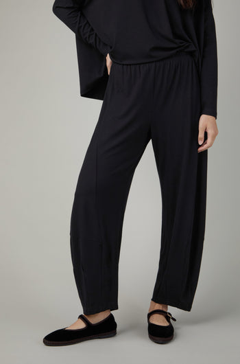 A person wearing Fluid Crepe Black Bubble Trousers, a long-sleeve black top, and black flat shoes stands with one hand on their hip against a plain background.