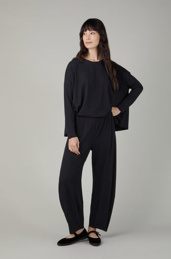 Woman stands against a plain background wearing Fluid Crepe Black Bubble Trousers, paired with a loose black long-sleeve top and black flat shoes.