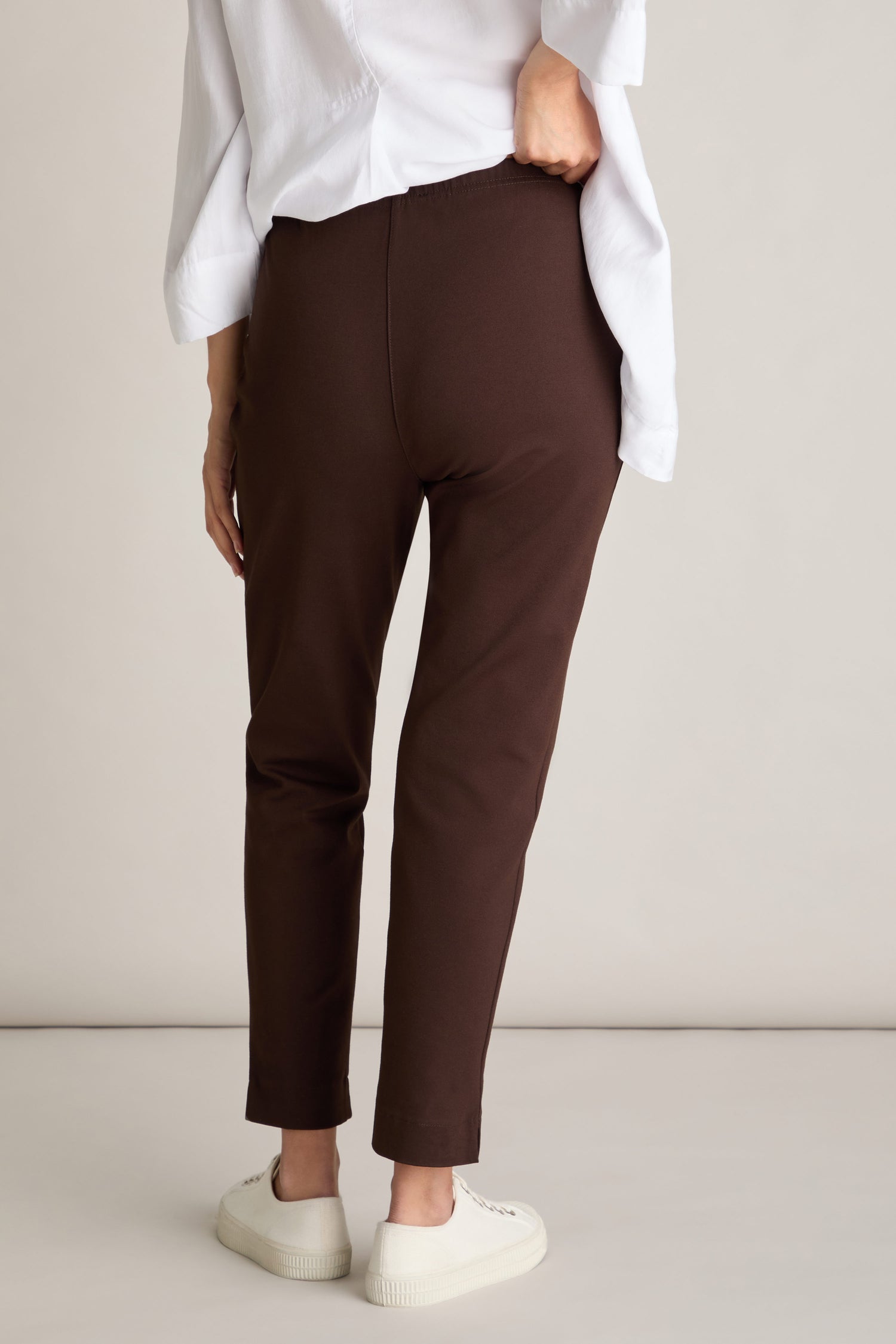 A person is shown from the back wearing a white top, brown pants, and white sneakers. The plain and neutral background highlights the Ponte Slim Leg Trouser, featuring an elasticated waistband for added comfort.