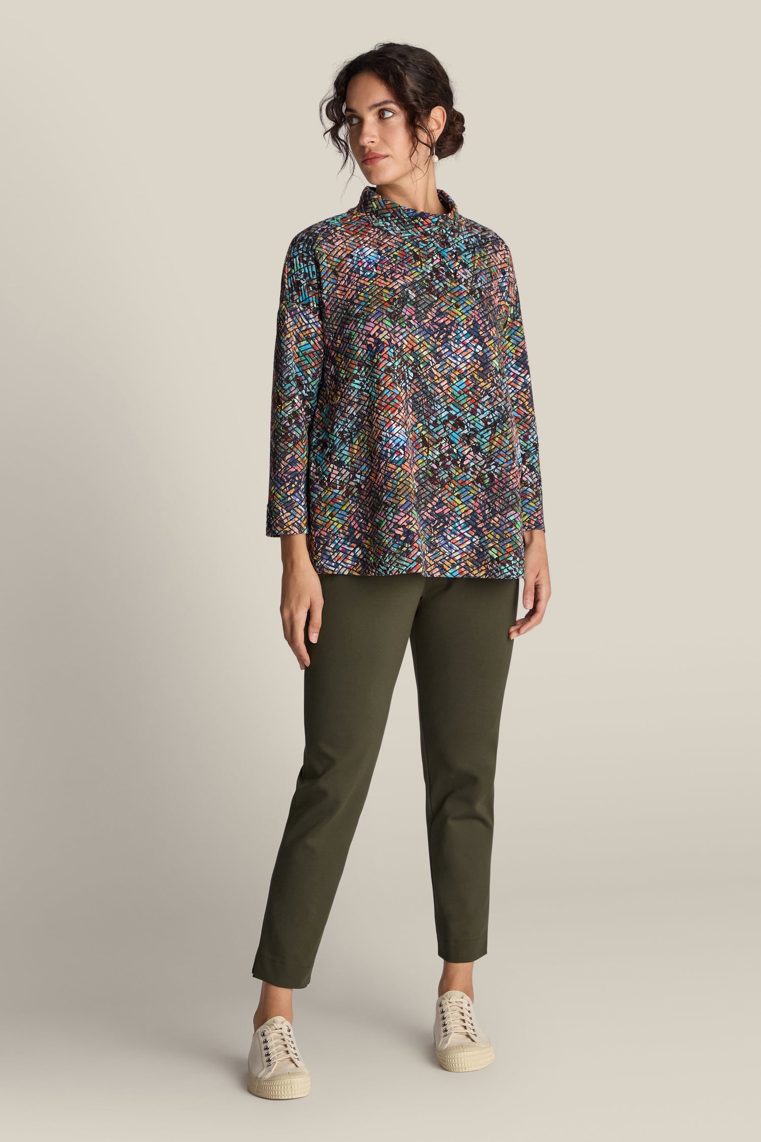 A woman stands against a beige background, wearing a colorful patterned top with long sleeves, olive green **Ponte Slim Leg Trousers** featuring an elasticated waistband for that tailored touch, and beige sneakers.