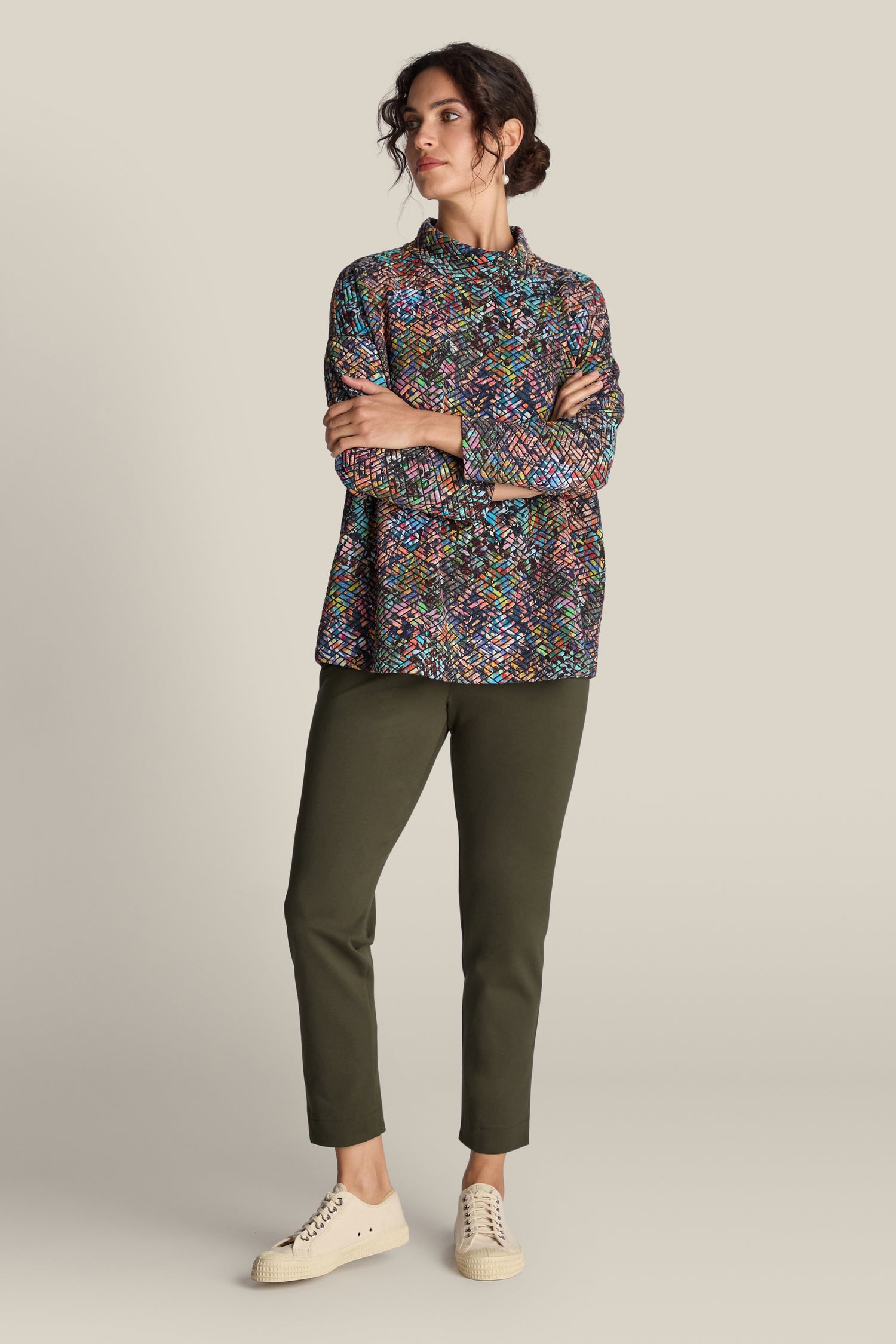 Person in a multi-colored, long-sleeve top and olive green **Ponte Slim Leg Trouser** standing with arms crossed. Cream-colored sneakers complete the look. Neutral background.