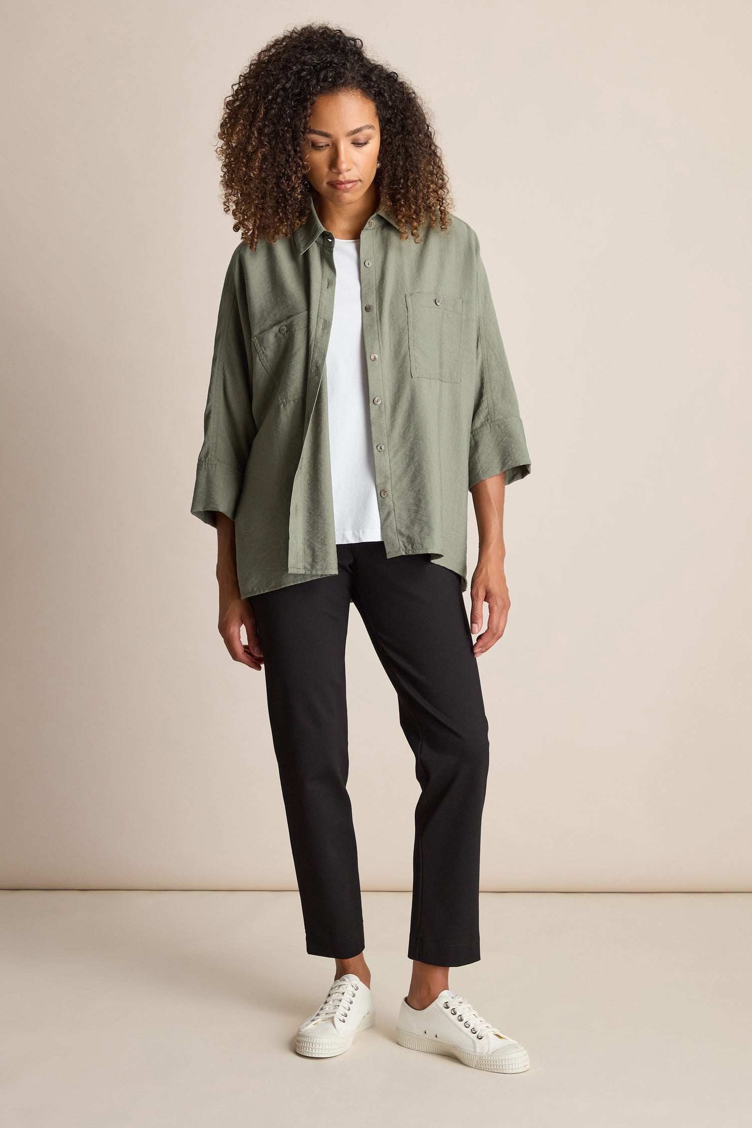 A woman with curly hair is wearing a green button-up shirt, a white t-shirt, black Ponte Slim Leg Trousers with an elasticated waist, and white sneakers. She is standing against a plain light-colored background.