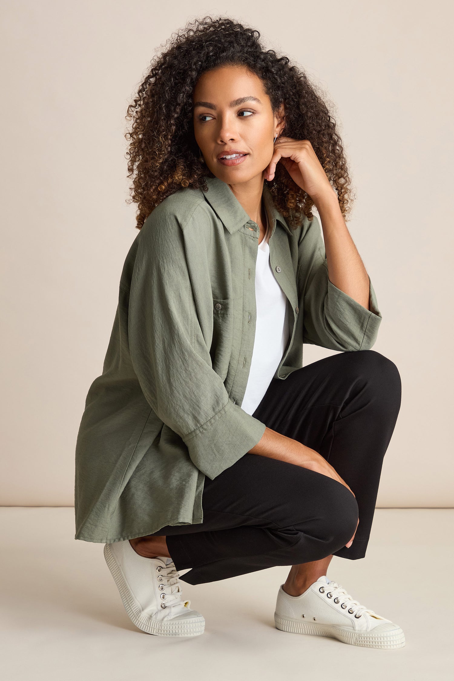 A person with curly hair crouches while looking to the side, wearing a green button-up shirt, Ponte Slim Leg Trousers, and white sneakers.