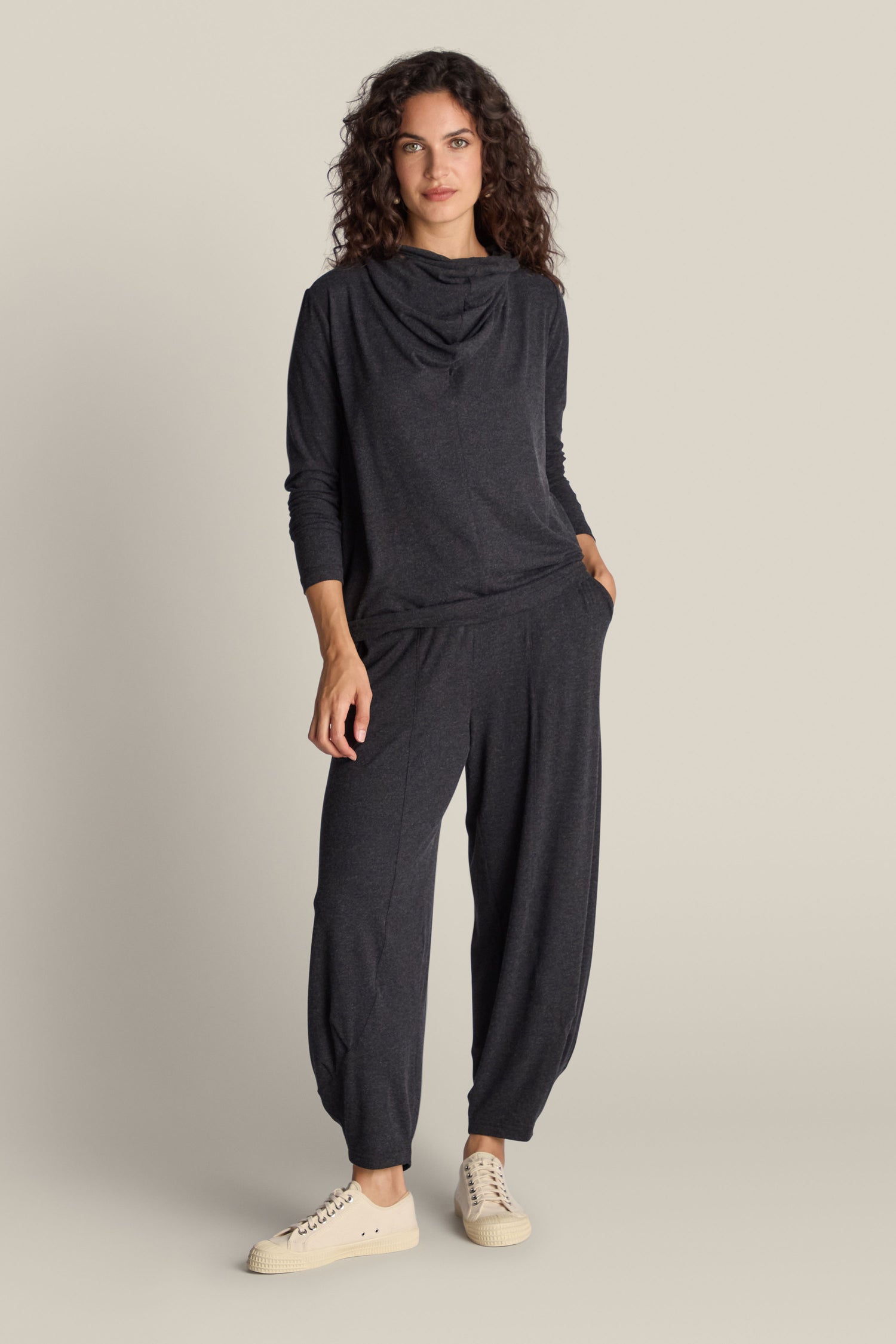 A person with long curly hair stands against a plain background, wearing a loose, dark grey top and matching Soft Viscose Jersey Bubble Trouser. Hands in pockets and white sneakers complete their flattering silhouette, embodying the essence of luxury loungewear.