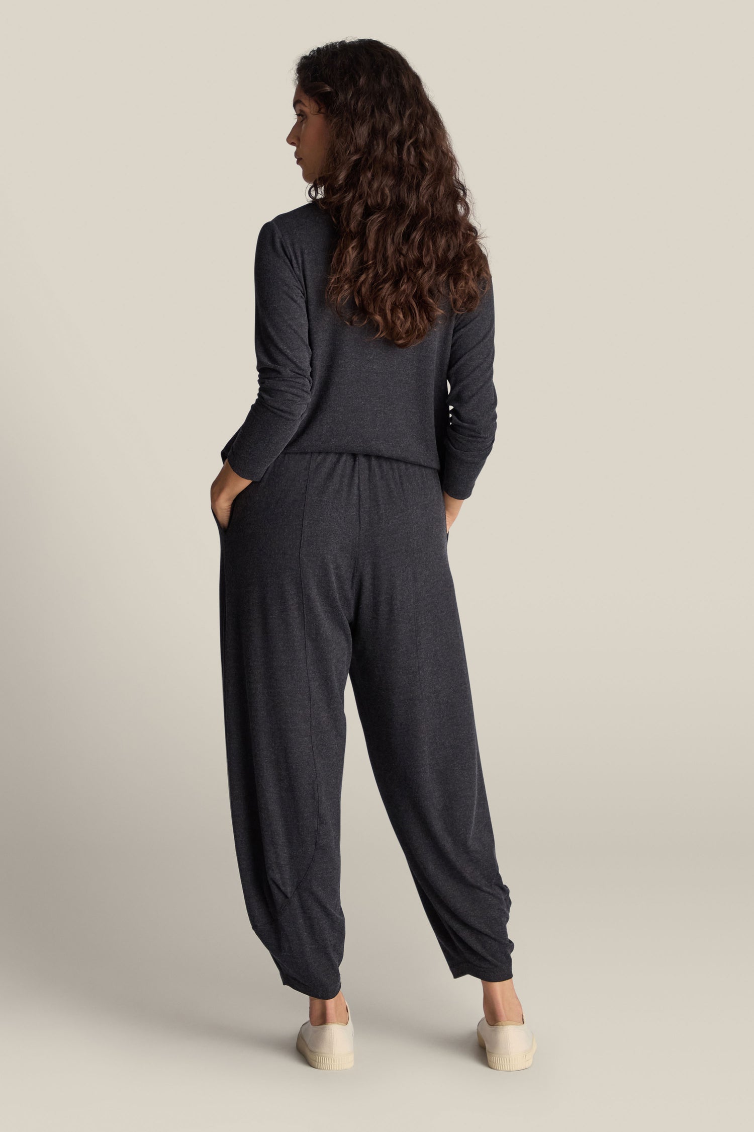 A person with long, curly hair stands with hands in pockets, wearing dark gray Soft Viscose Jersey Bubble Trousers and white slip-on shoes, presenting a flattering silhouette while facing away from the camera.