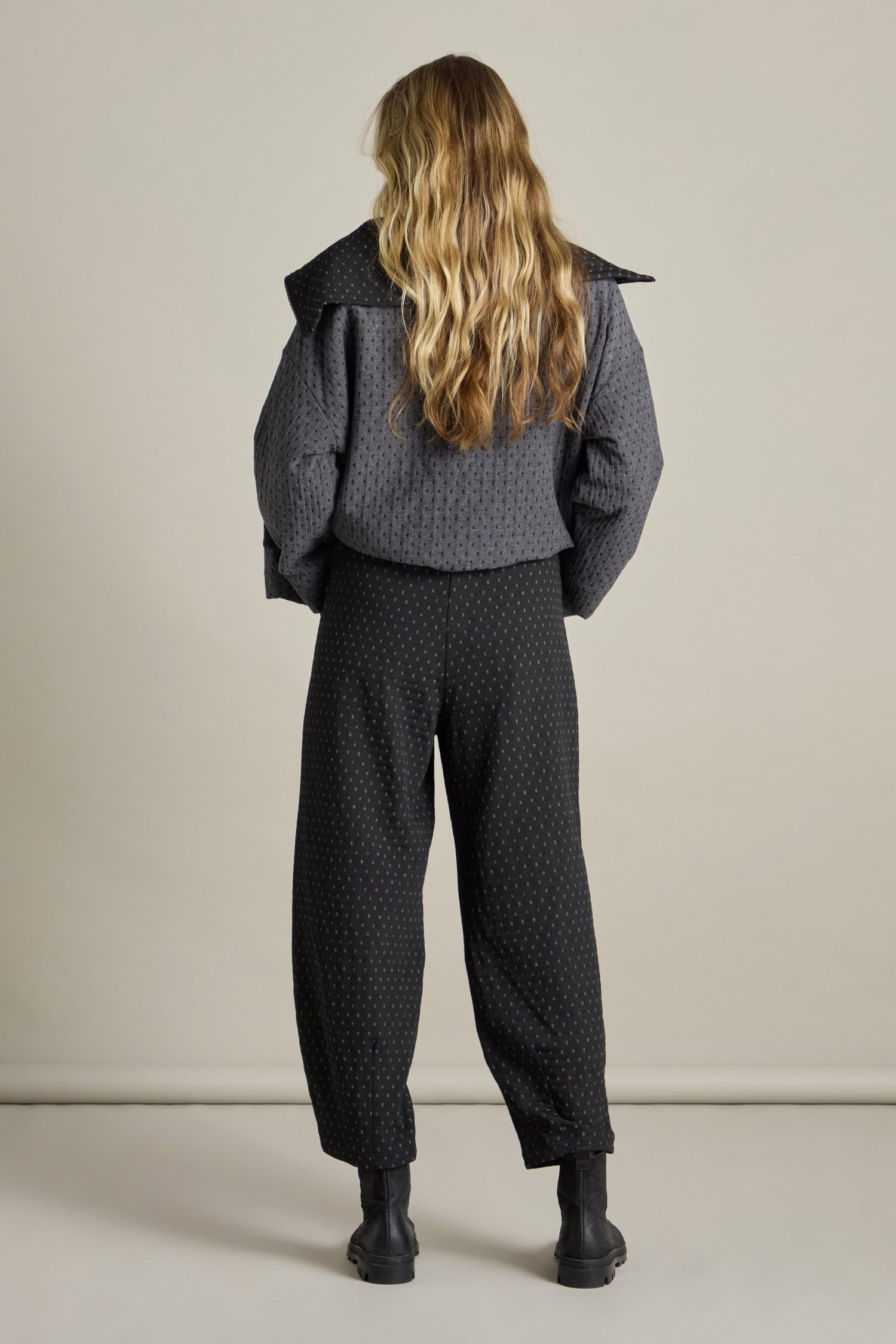 A person with long, wavy hair stands facing away from the camera against a plain background, wearing a textured dark jacket made of double-faced jacquard fabric and stylishly cropped Square Jacquard Bubble Trousers.