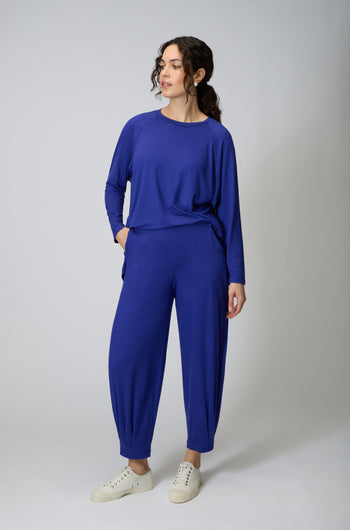 A woman stands in a studio wearing Soft Viscose Jersey Bubble Trousers and a matching long-sleeve top with white sneakers, looking to the side with one hand in her pocket.