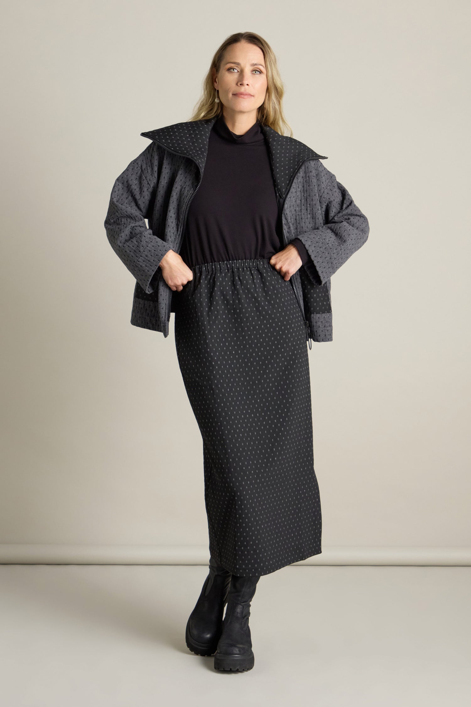 A woman stands in front of a plain background wearing a dark top, a gray jacket with a wide collar, and black boots. Her long Square Jacquard Tube Skirt features a tube silhouette and is crafted from double-faced jacquard fabric, adding an elegant touch to her ensemble.