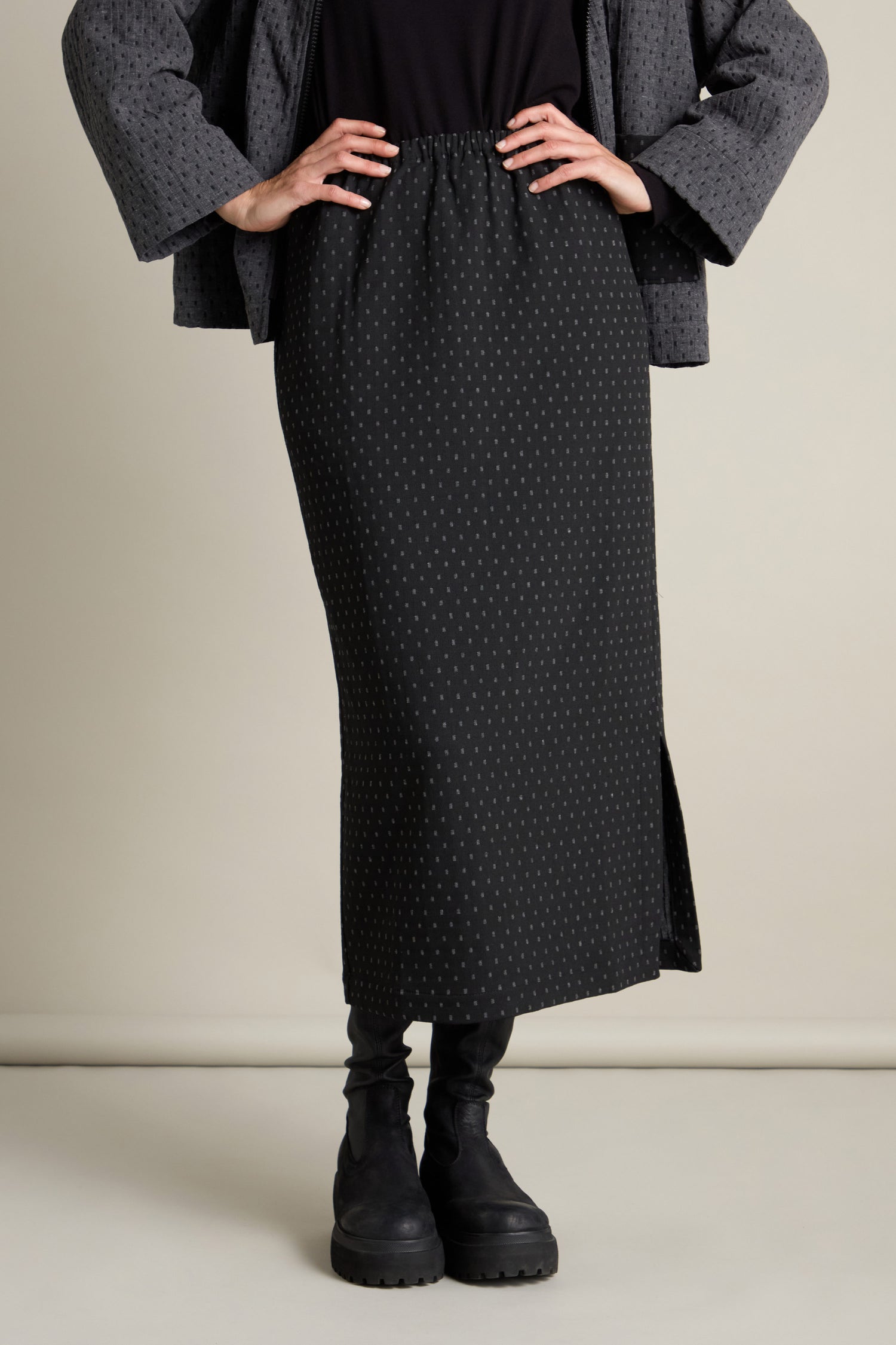 The individual stands confidently in a Square Jacquard Tube Skirt, crafted from luxurious double-faced jacquard fabric. The skirt, accentuated by a stylish side slit, pairs seamlessly with a dark polka dot jacket and black boots. With hands placed on their hips, the outfit highlights their poised stance.