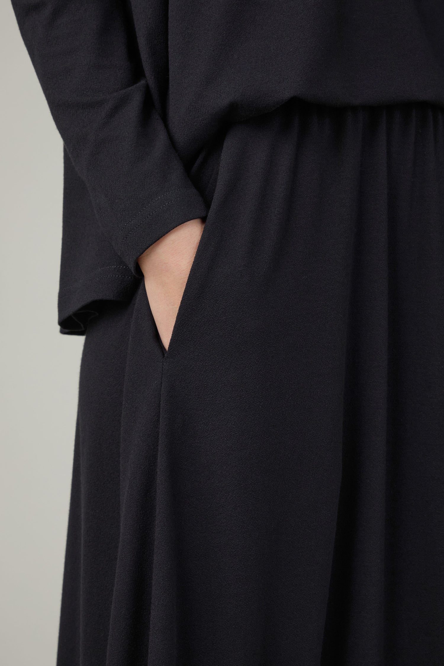 A person wears a loose, long-sleeved black top and places their right hand in the side pocket of the chic Fluid Crepe Black Bubble Skirt.