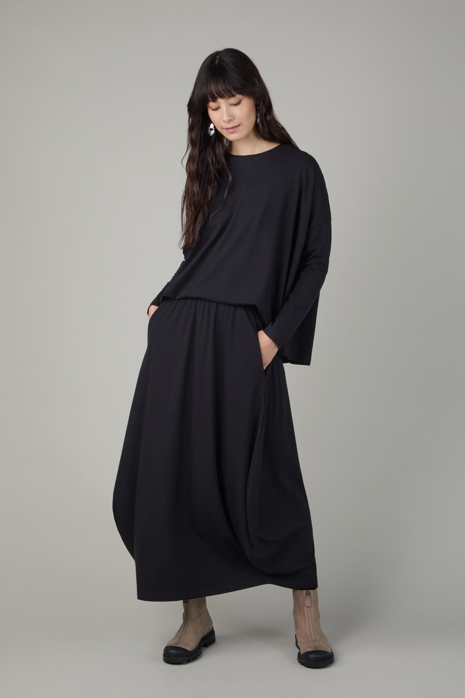 A woman stands against a plain background wearing a loose black top and the Fluid Crepe Black Bubble Skirt with hands in pockets, paired with taupe ankle boots.