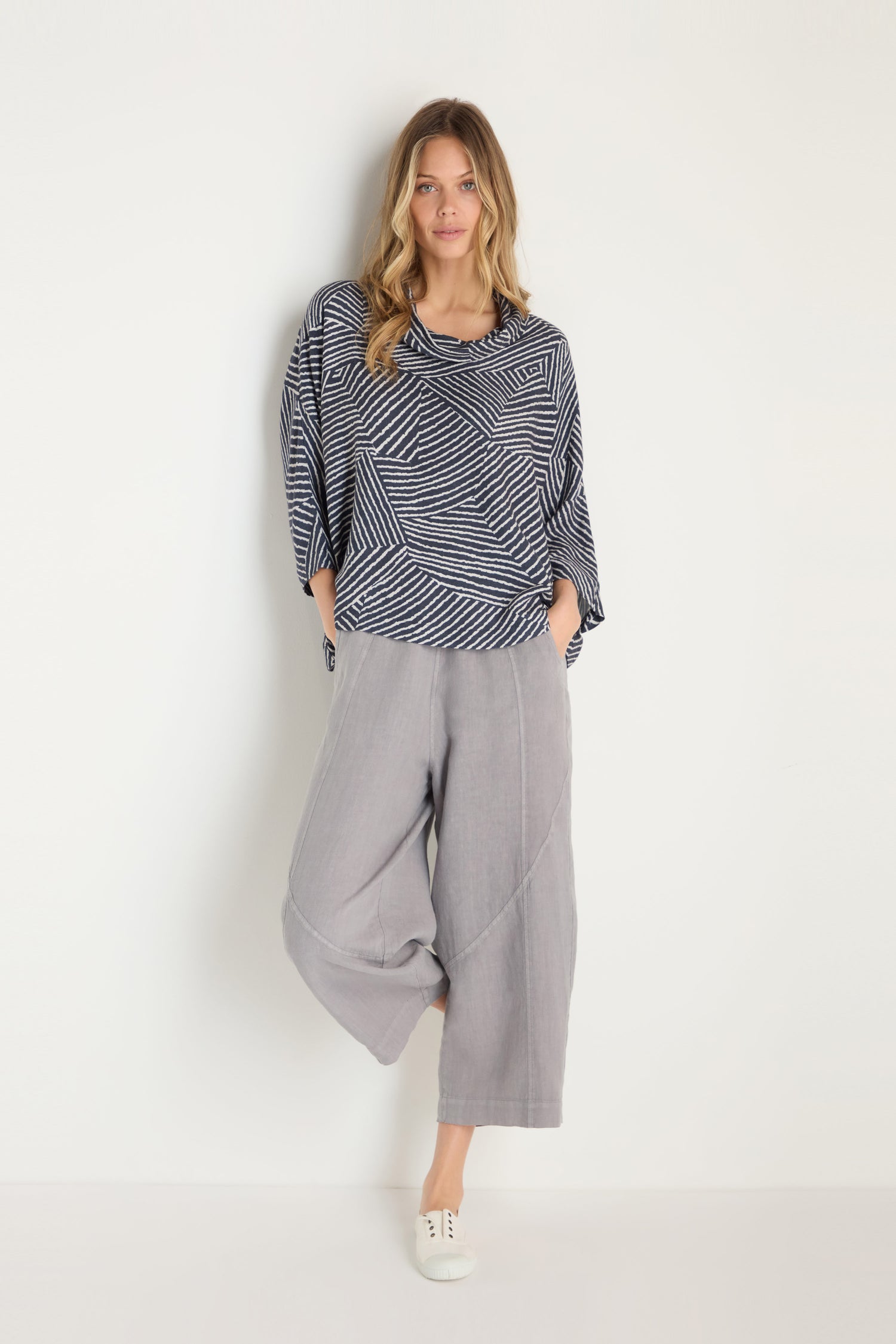 A woman stands against a white wall wearing the Linear Prism Lines Cowl Neck Top, wide-leg gray pants, and white sneakers, with her hands in her pockets.