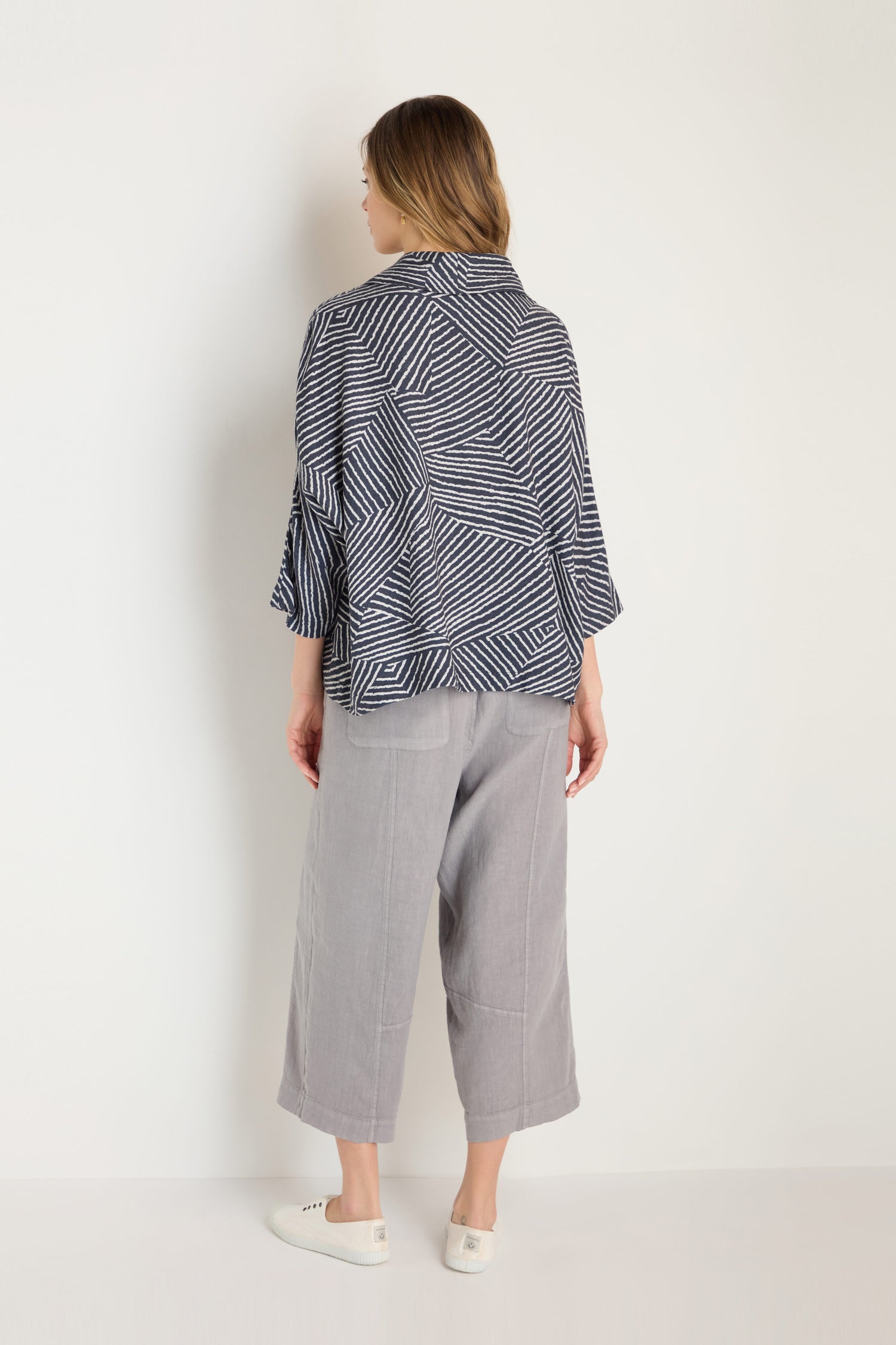 A woman stands facing away, wearing the Linear Prism Lines Cowl Neck Top in navy and white geometric print, paired with light gray wide-leg pants and white slip-on shoes, all set against a plain white background.