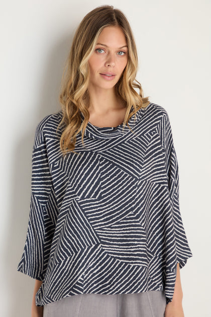 Woman with wavy blond hair wears the Linear Prism Lines Cowl Neck Top, a loose-fitting navy and white geometric print piece crafted from soft brushed viscose, standing against a plain light background.