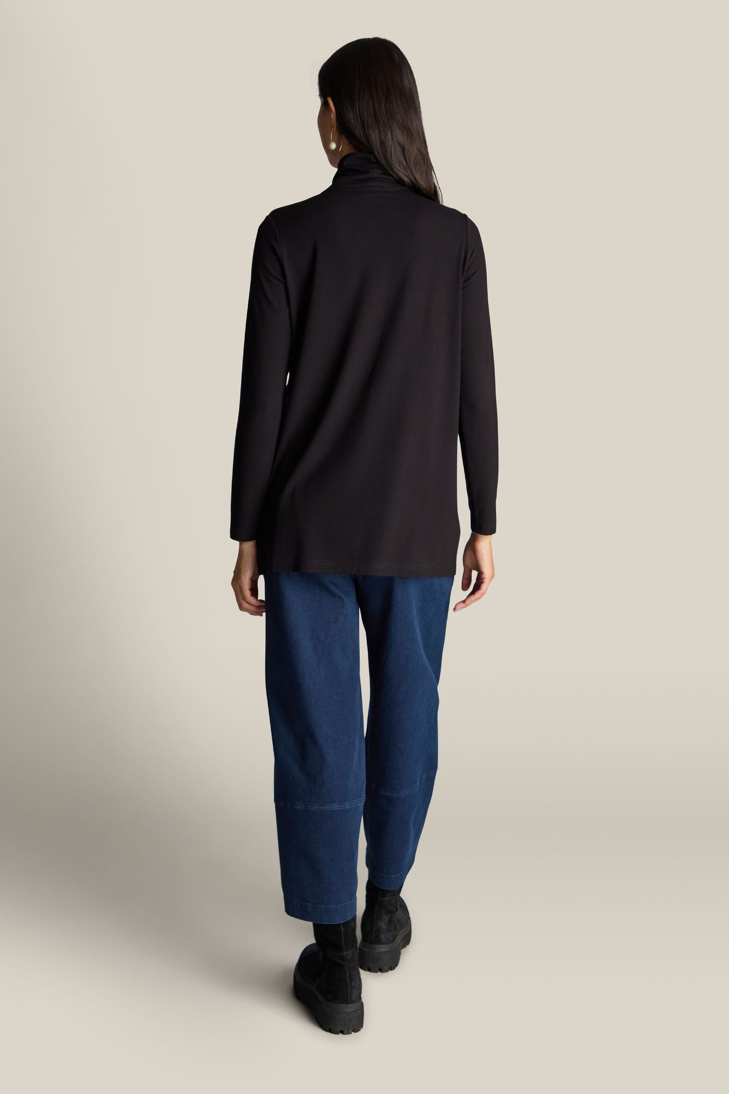 Rear view of a person with long hair in a black Fluid Crepe Flared Polo Neck, blue wide-leg jeans, and black shoes, standing against a plain, neutral background—a perfect addition to an elegant wardrobe.