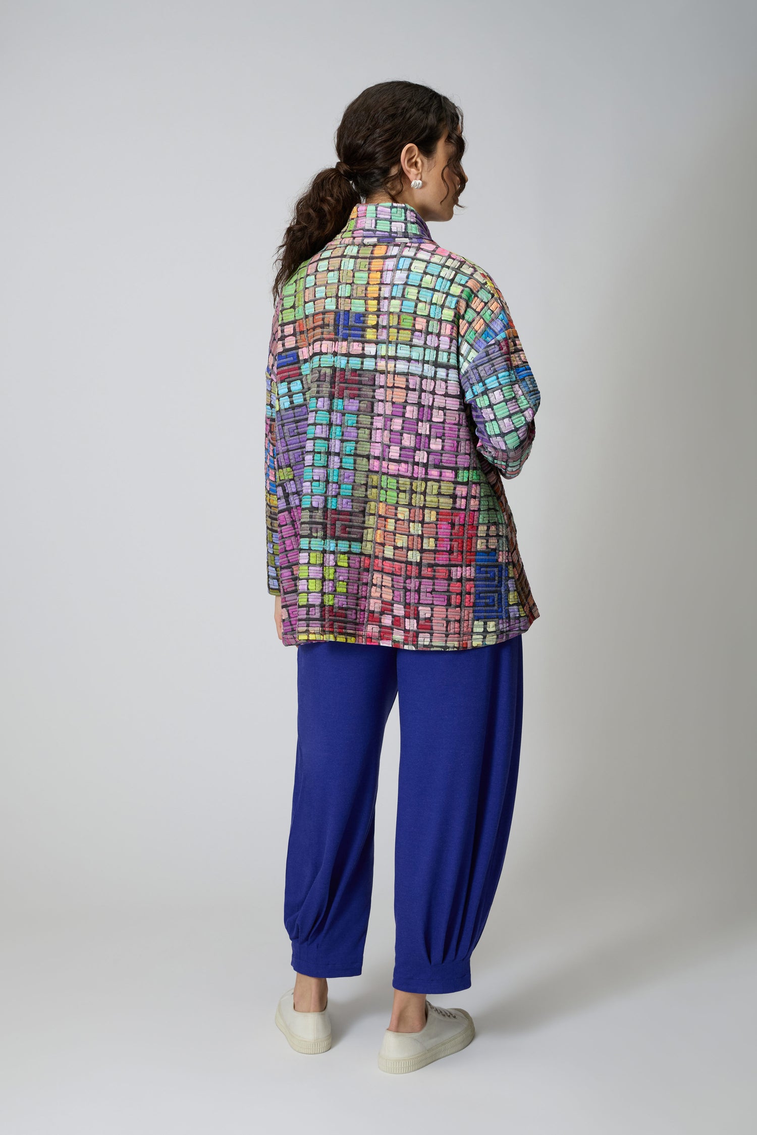 A person stands facing away in a plain studio, wearing the Coloured Glass Tiles Jersey Tunic—a colourful, grid-patterned piece—paired with blue pants, white shoes, and hoop earrings.