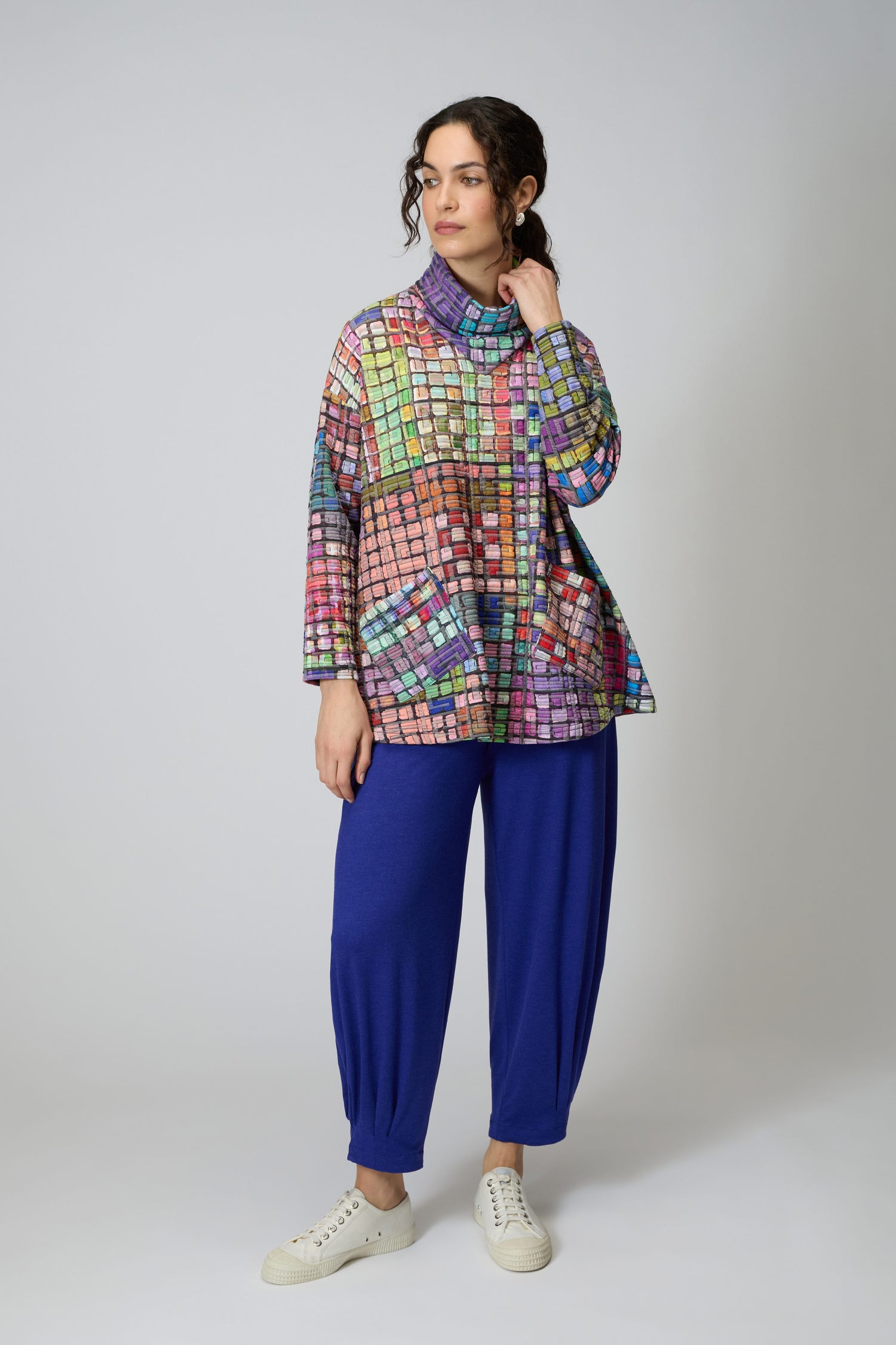 A woman wears the Coloured Glass Tiles Jersey Tunic with blue pants and white sneakers, standing against a plain light gray background.