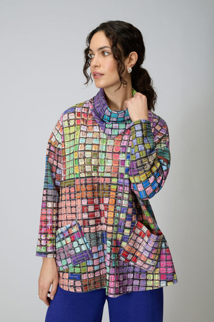 A woman wears the Coloured Glass Tiles Jersey Tunic—an oversized, grid-patterned design with large pockets—paired with blue pants against a plain light background.