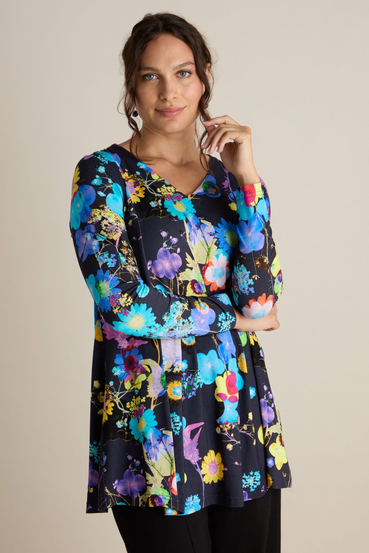 A person wearing a long-sleeved, vibrant Blue Bouquet Jersey Top and black pants is standing against a plain background, touching their chin with one hand and looking at the camera, highlighting this elegant addition to any autumn wardrobe.