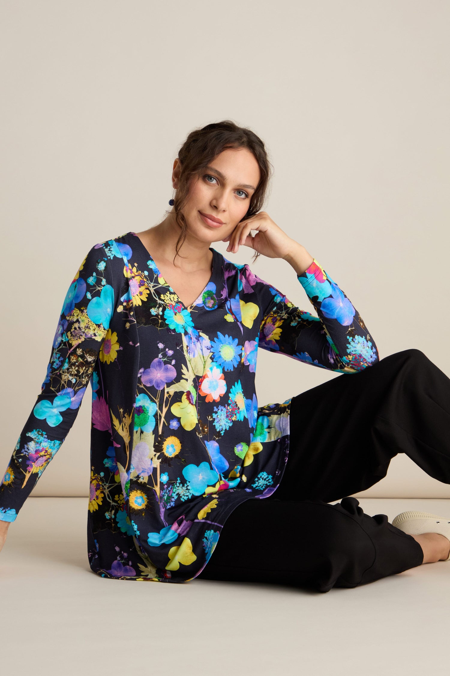 A person is seated on the floor against a plain background, wearing the Blue Bouquet Jersey Top with its colorful floral pattern and long sleeves, paired with black pants—perfect for an autumn wardrobe.