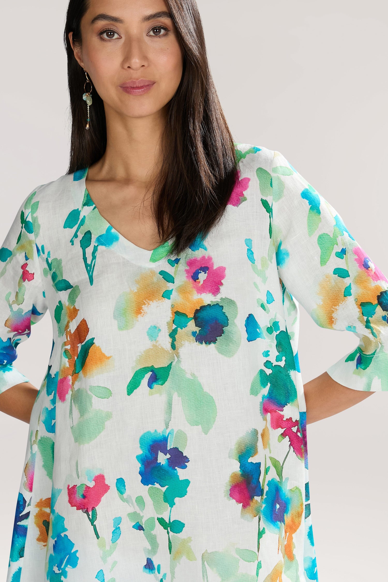 A woman wearing a Watercolour Foxglove Tunic stands against a plain background, looking at the camera. The Watercolour Foxglove Tunic features a colorful floral foxglove motif on pure linen. She has long dark hair and is accessorized with dangly earrings, adding an elegant touch to her look.