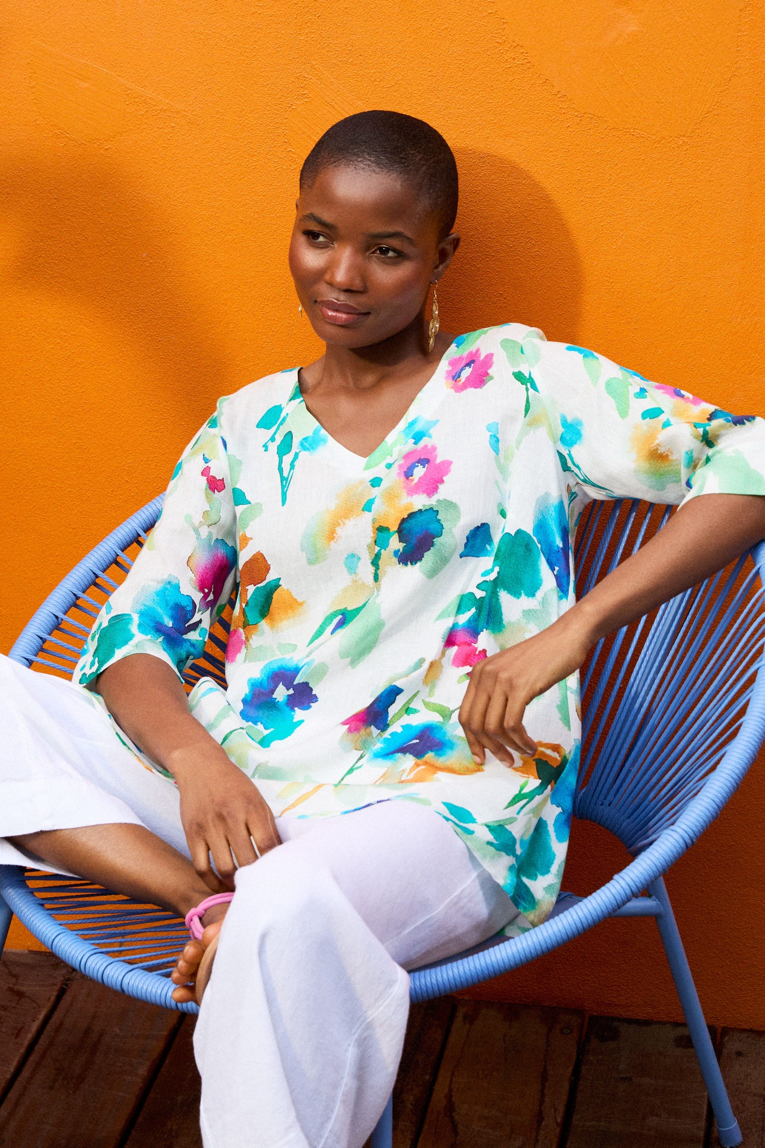 A person with short hair sits on a blue chair against an orange wall. They wear a Watercolour Foxglove Tunic and white pants, with one arm resting on the chair's armrest.