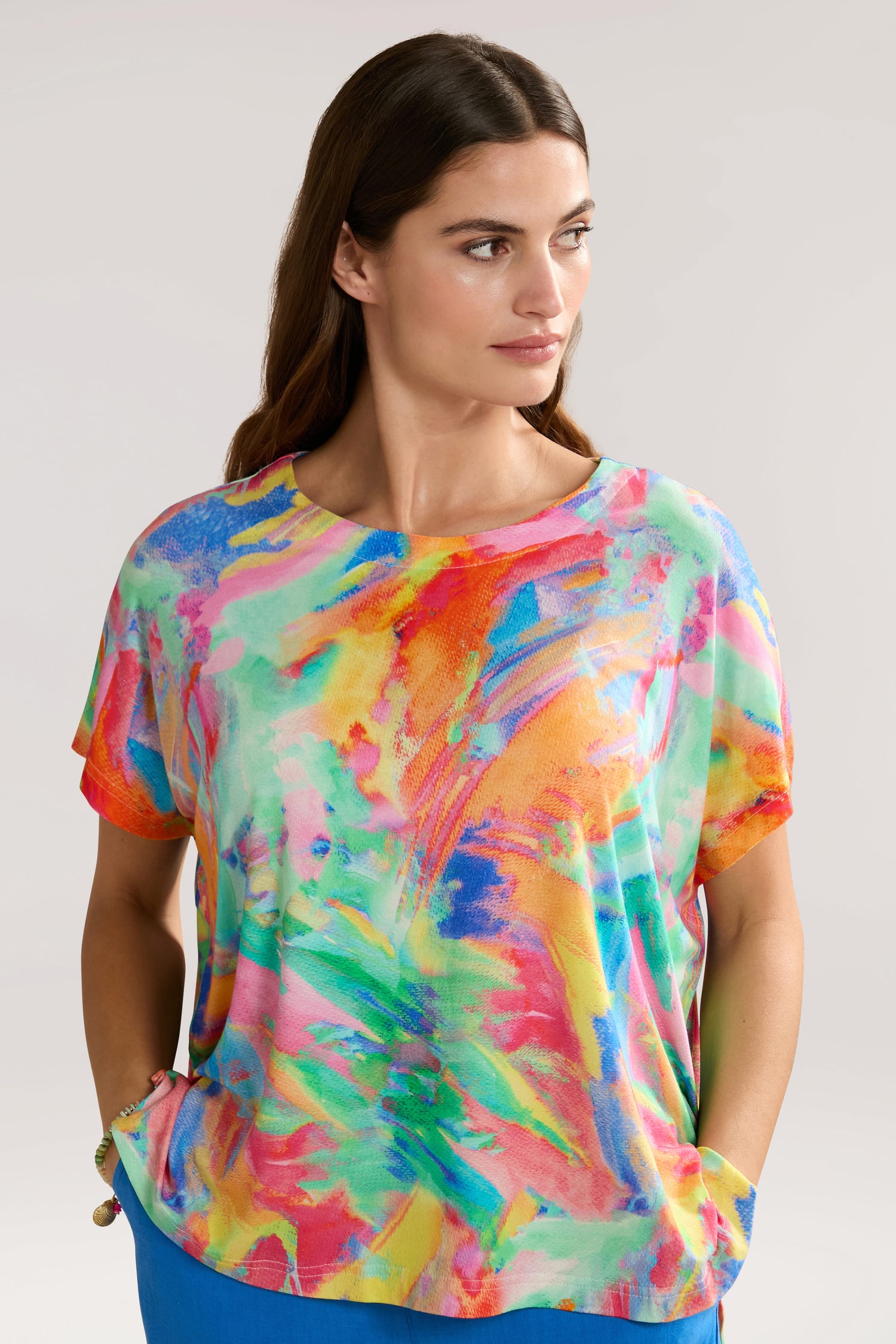 A woman in a colorful Vibrant Watercolour Jersey Top with abstract patterns stands with her hands in her pockets, looking to the side. She has long brown hair and a neutral expression. The background is plain light gray, complementing her elegant style crafted from sustainable LENZING™ Viscose ECOVERO™.