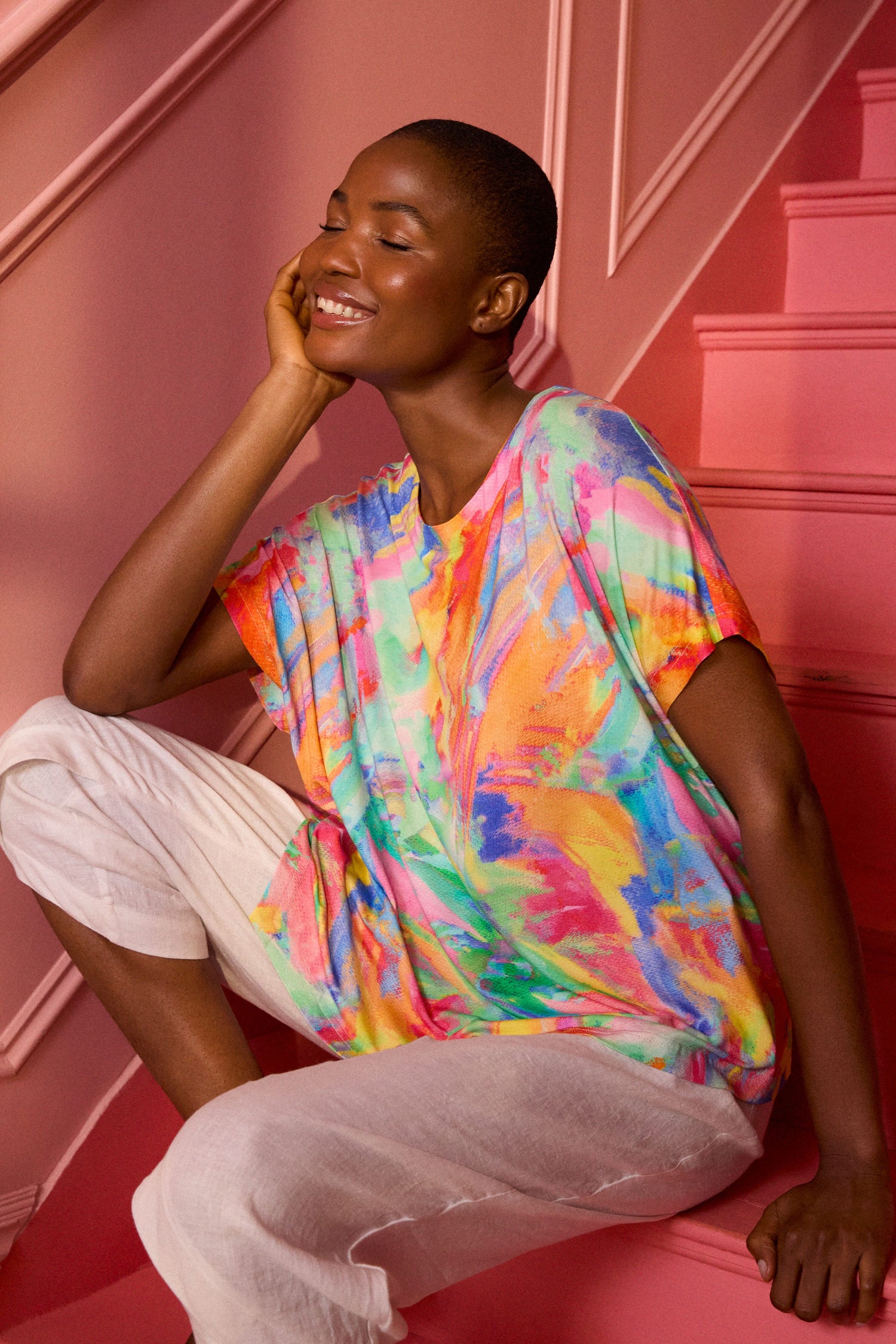 A person wearing a Vibrant Watercolour Jersey Top and white pants sits on pink stairs, smiling with their eyes closed, resting their head on their hand.