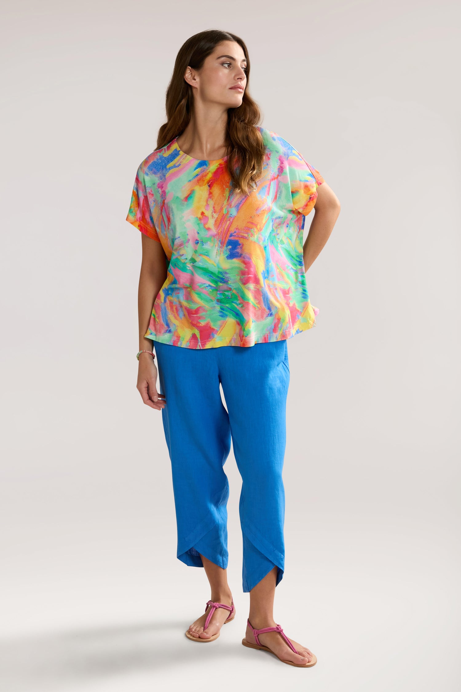 A woman stands against a plain background wearing an elegant Vibrant Watercolour Jersey Top made from LENZING™ Viscose ECOVERO™, blue pants, and pink sandals, looking to her right.