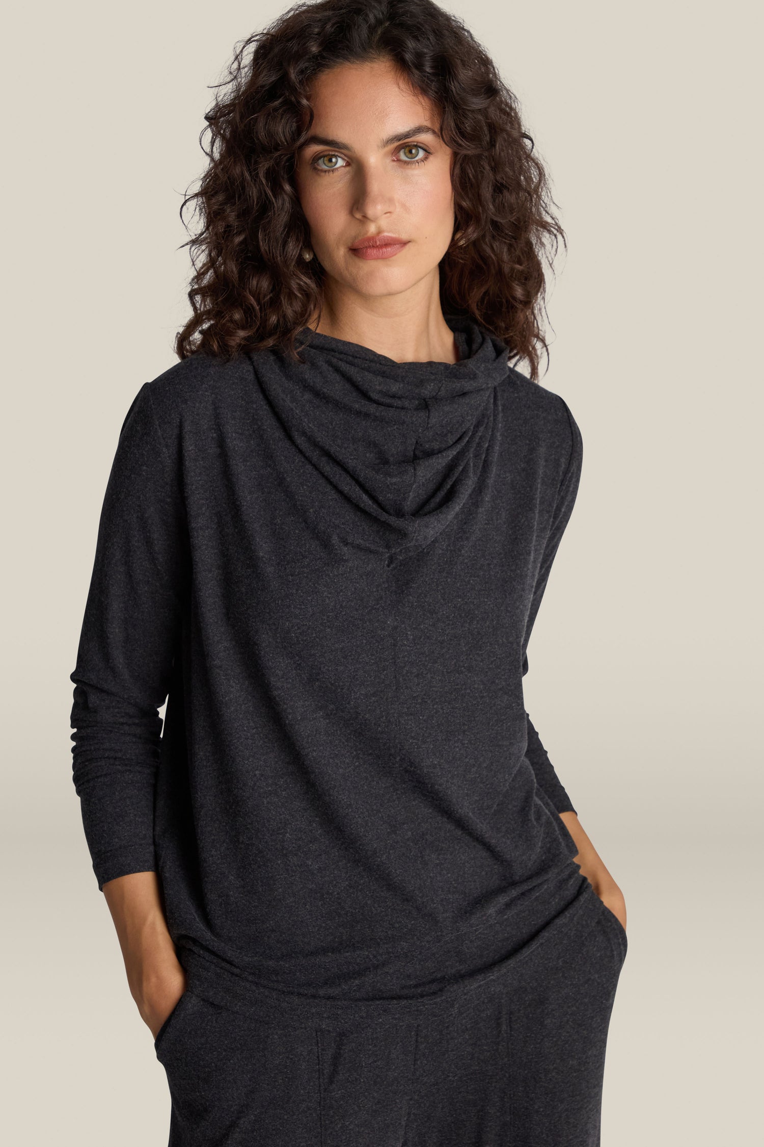 A person with curly hair is wearing a Soft Viscose Jersey Cowl Neck Top in dark gray, featuring long sleeves and their hands in pockets. They stand against a plain background, exuding both elegance and comfort.