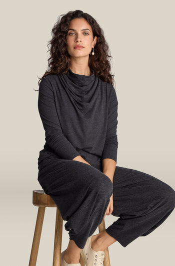 A person with curly hair sits on a wooden stool wearing an elegant dark grey Soft Viscose Jersey Cowl Neck Top and matching pants, paired with light-colored shoes.