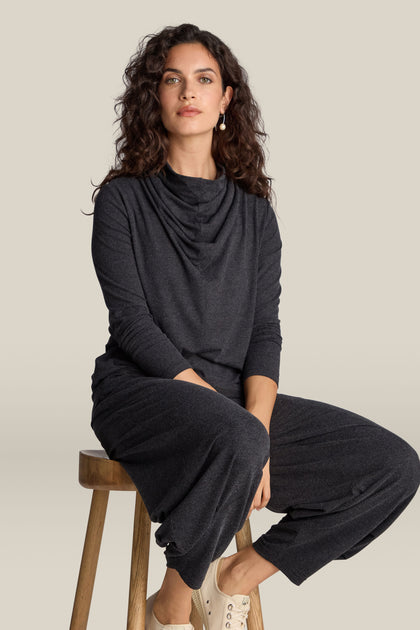 A person with curly hair sits on a wooden stool wearing an elegant dark grey Soft Viscose Jersey Cowl Neck Top and matching pants, paired with light-colored shoes.