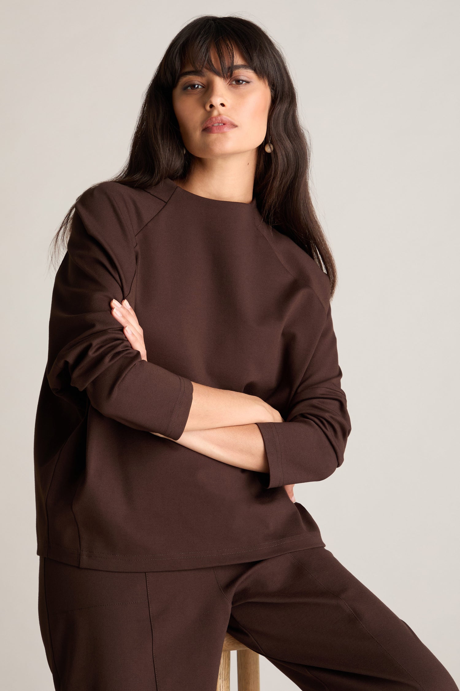 A woman with long dark hair, wearing a coordinating brown Ponte Boxy Top and matching pants, sits with her arms crossed, looking straight ahead and exuding a casual chic look.