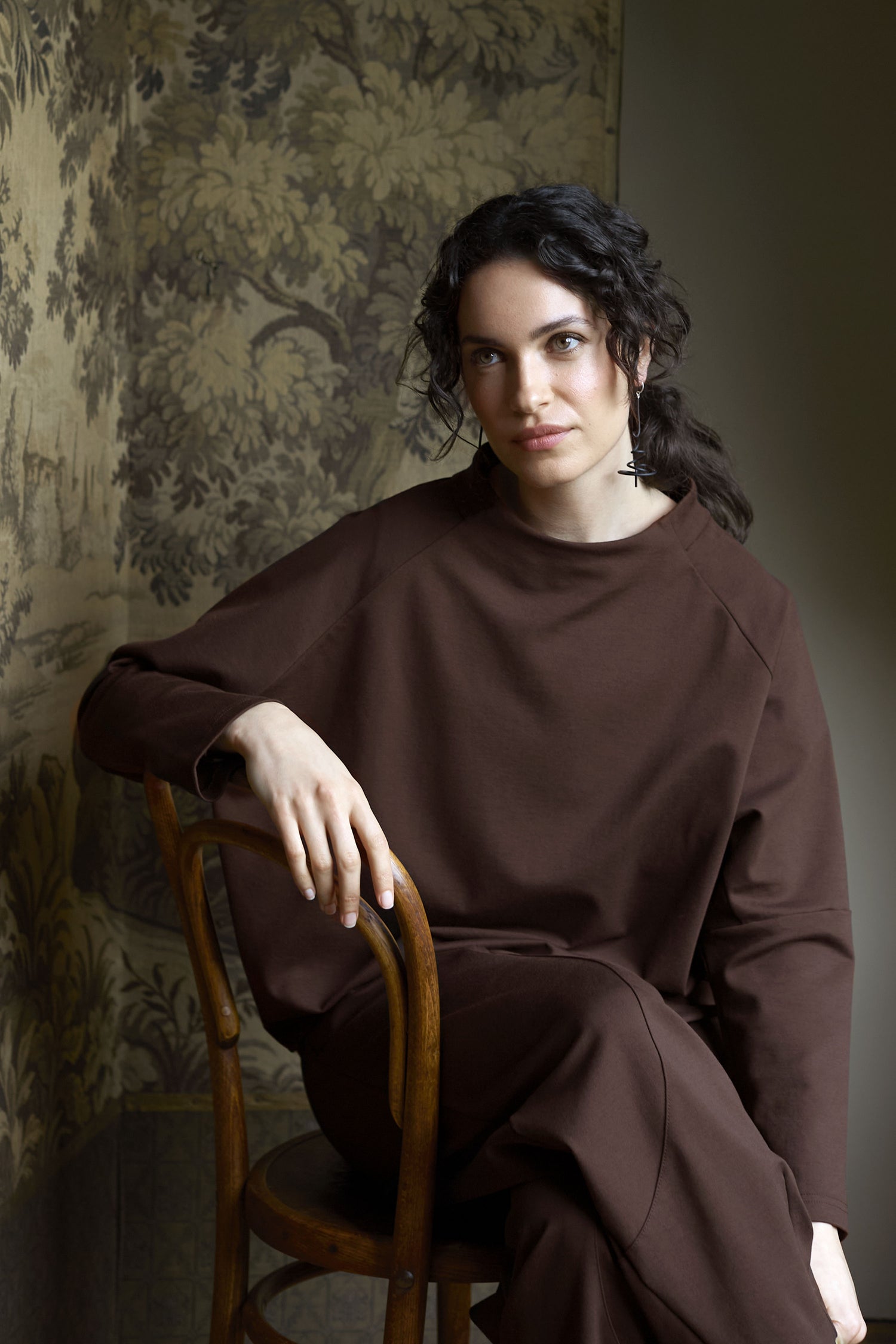 A person with dark hair sits on a wooden chair, wearing the Ponte Boxy Top in brown, which accentuates their casual chic style. The background features a detailed, green-toned wall design, creating a striking linear silhouette.